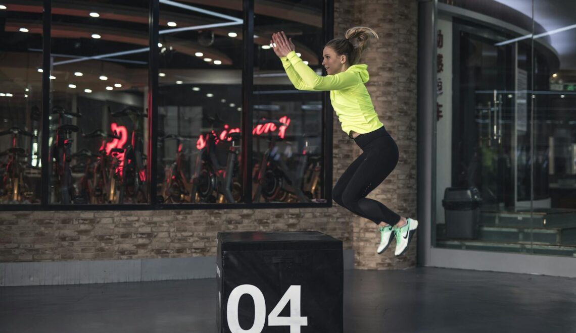 Fit young woman in sports wear executing a box jump indoors, promoting fitness and health in a modern urban gym.